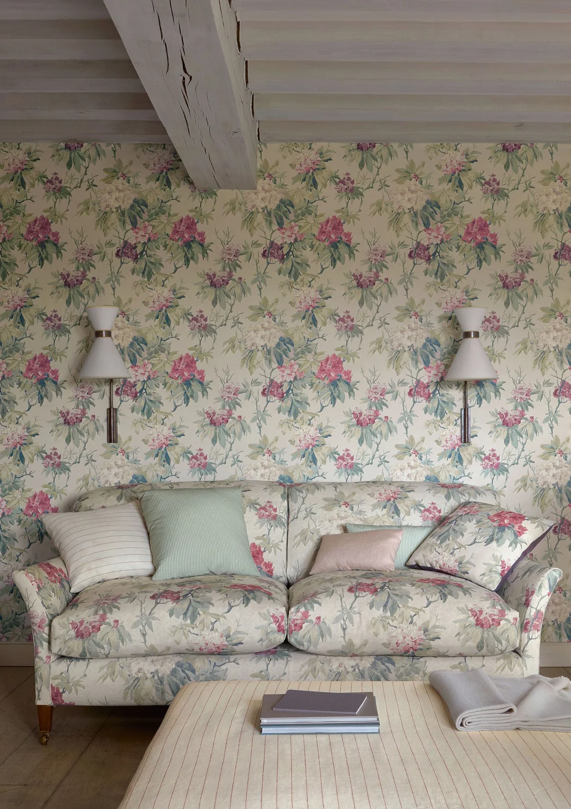 A floral sofa repeats the flower pattern on the wall behind it beneath white-painted beams.