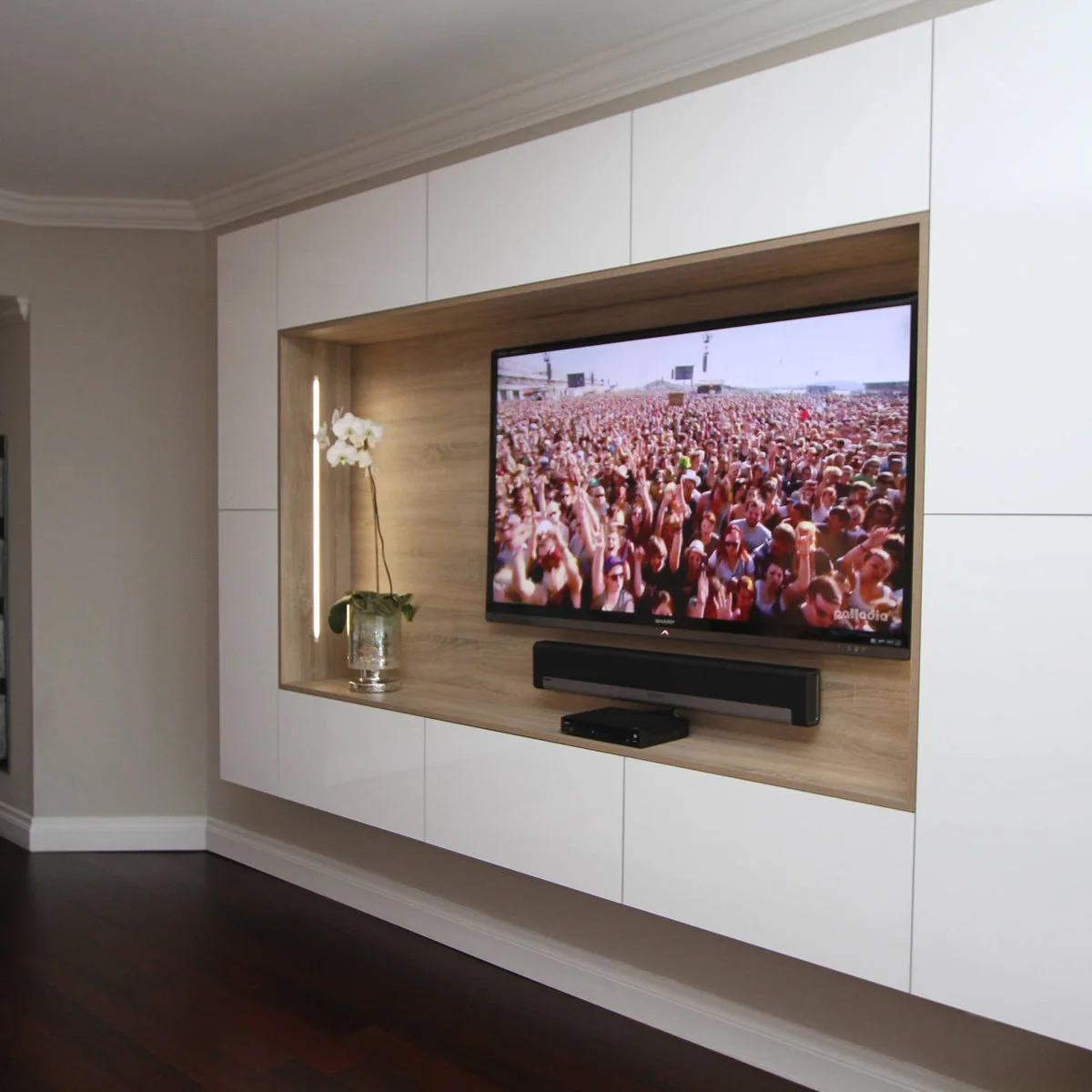 A television and soundbar occupy a wood-lined recess in white cabinetry, with one orchid placed at its left side.