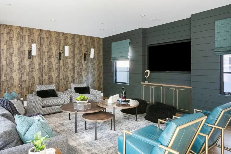Living room with dark horizontal-paneled television wall, low cabinet, teal chairs, gray sofa, round nesting tables, patterned wallpaper and two small window shades.