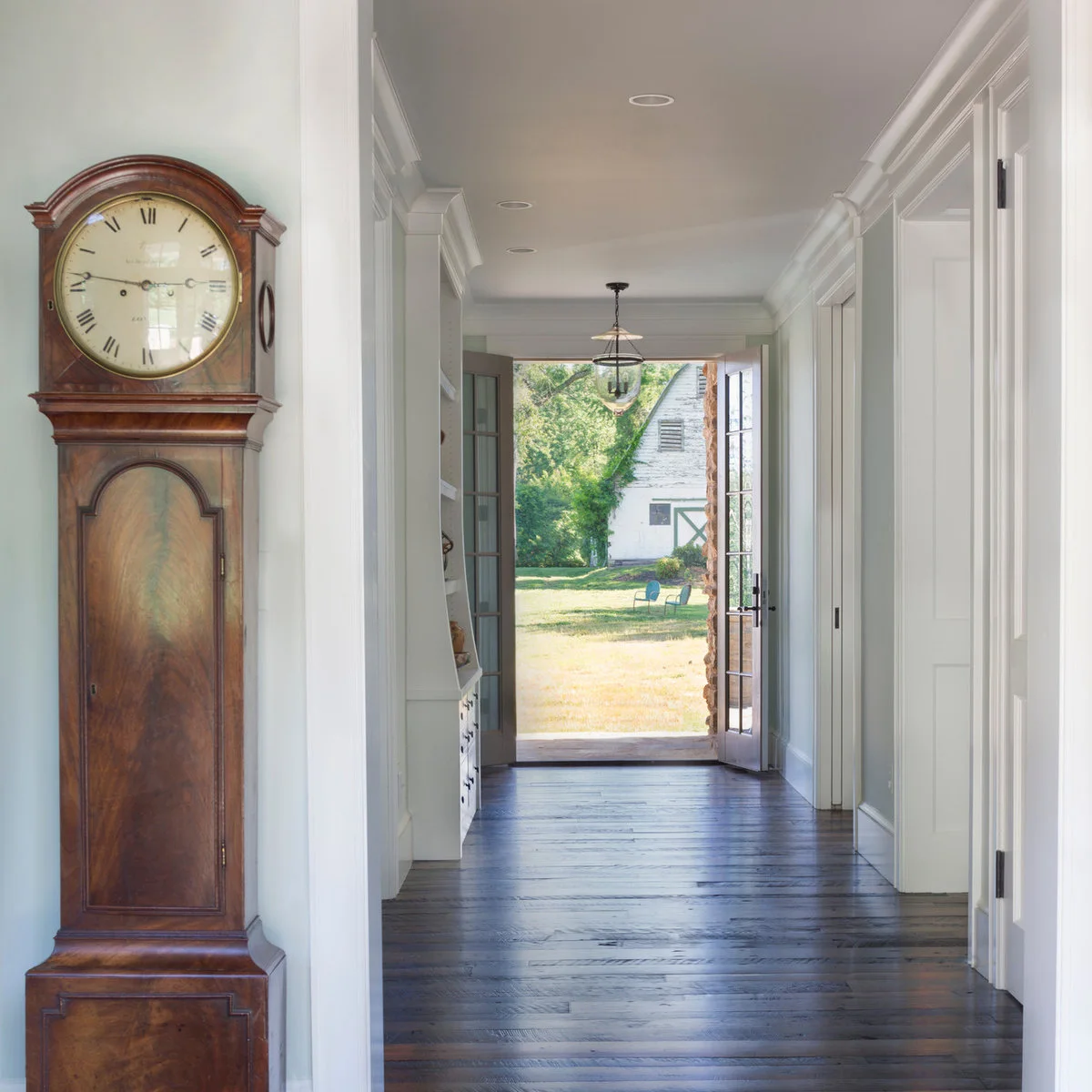 Pale entrance hall with dark wood flooring, a tall wooden clock and open glazed doors facing a garden.