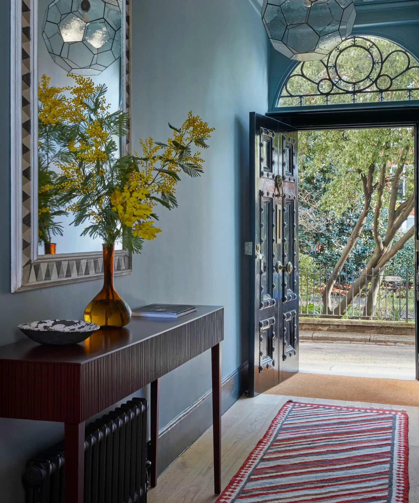 Small red and pale blue striped rug inside an open dark door, with blue walls, a ribbed console and mirrored wall