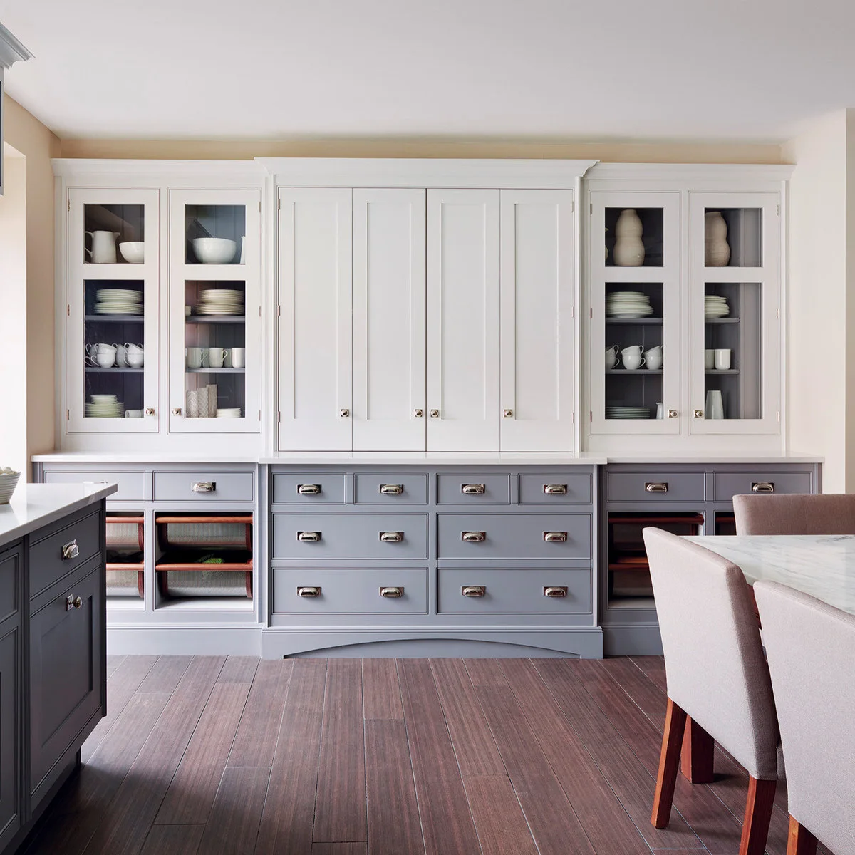 A tall pale cabinet display has gray lower drawers, dark glazed interiors and dusty rose dining chairs beside it.