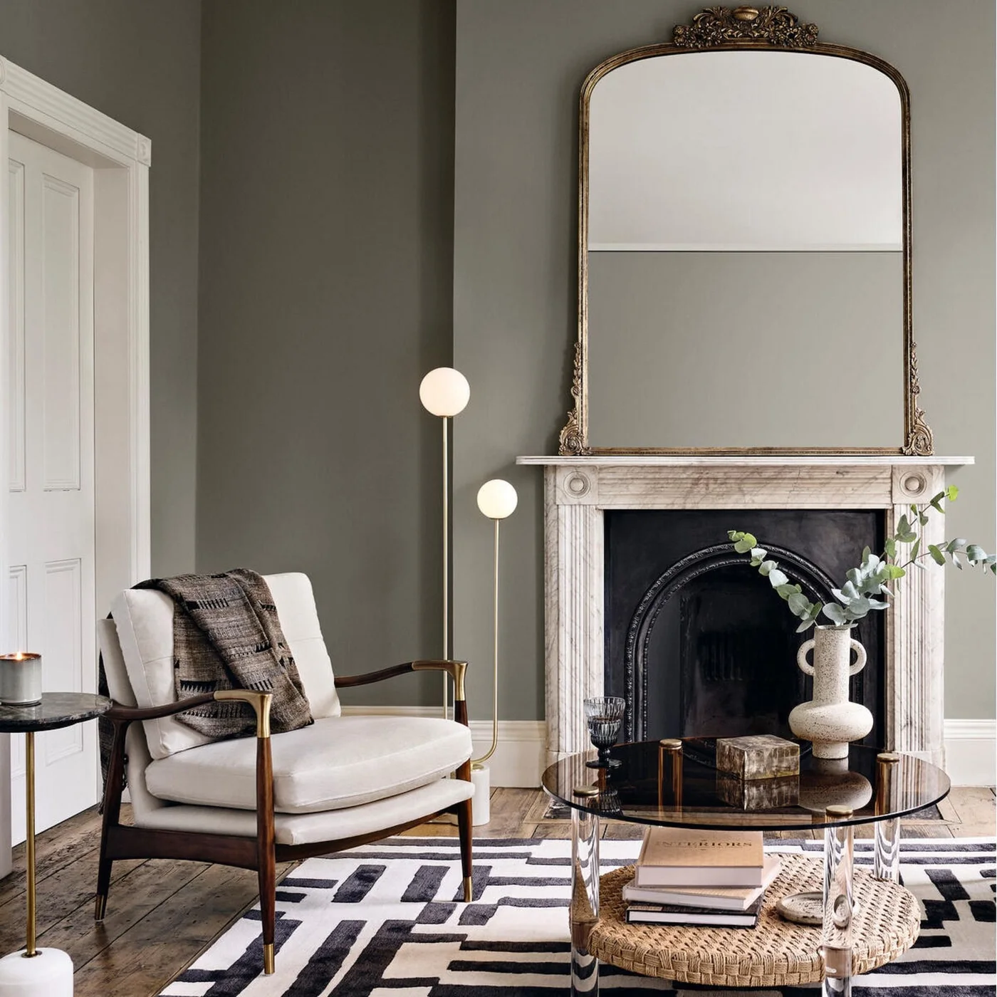 A pale veined marble surround and tall arched mirror sit against muted green walls beside a modern cream chair and round glass table.