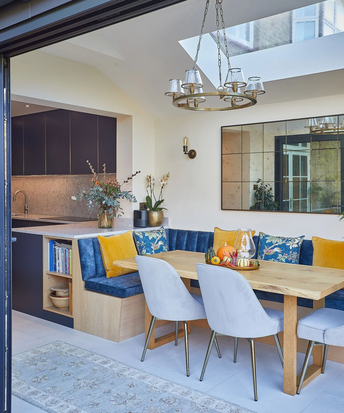 A blue upholstered corner bench with yellow cushions and an open end shelf sits beside a kitchen counter and wooden dining table