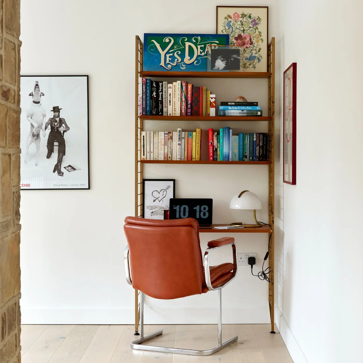 A narrow wood shelving unit includes a desk surface beneath books and artwork, with a brown upholstered metal-frame chair and a small white dome lamp.