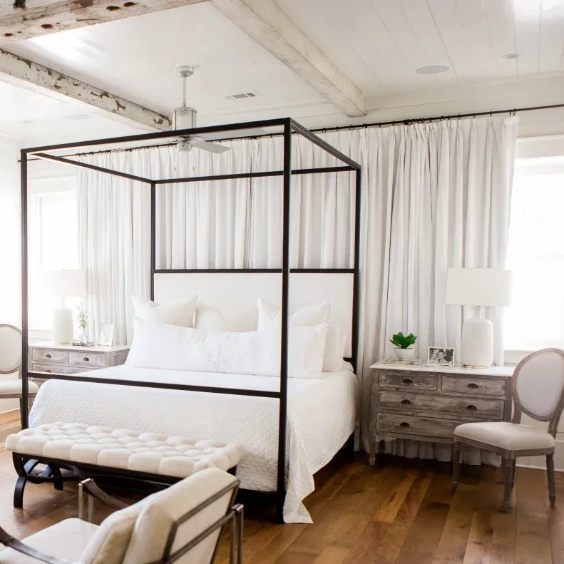 A dark canopy bed stands beneath pale exposed beams beside weathered timber drawer chests and white curtains.