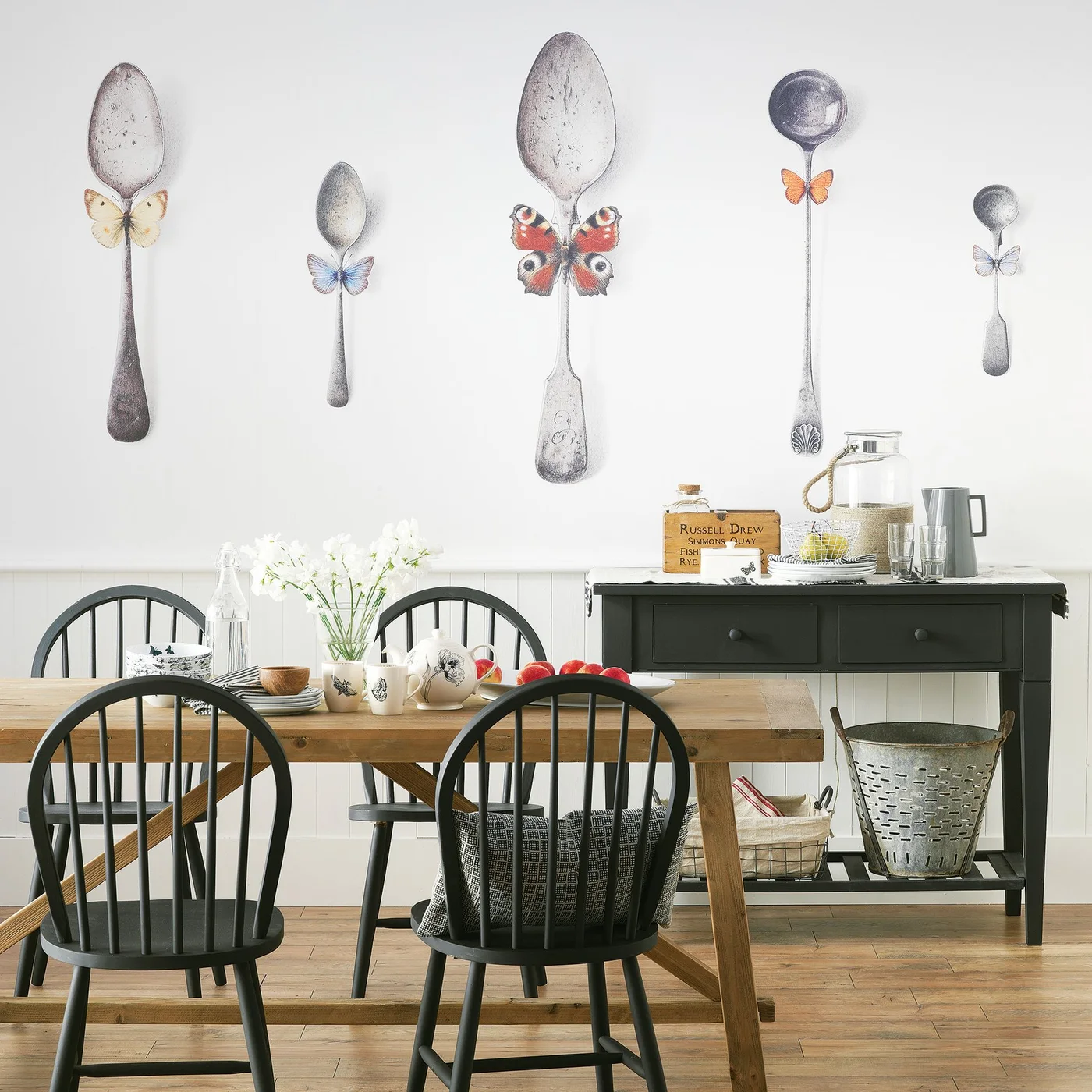 A black two-drawer console holds a large clear drink jar, tumblers and stacked plates, with baskets on its lower shelf in a dining room.