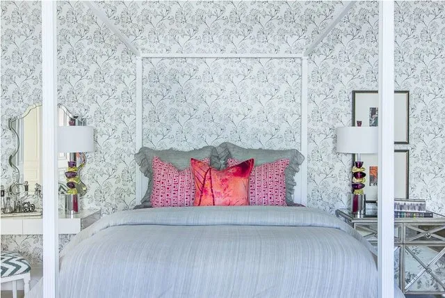Fine pale blue-gray branches cover the wall behind light bedding and bright pink cushions.