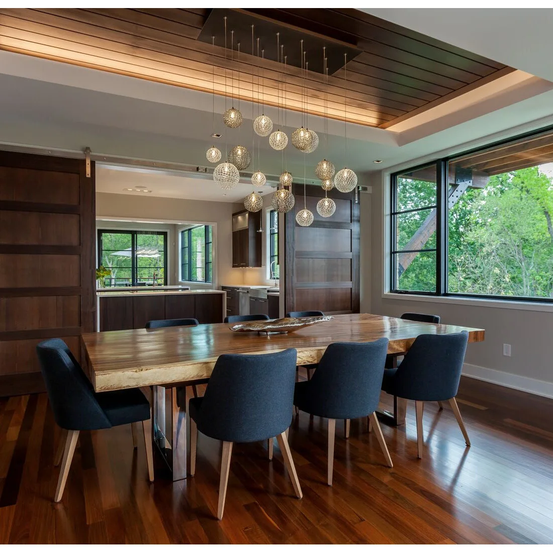 A wood dining table with an irregular edge stands beside blue upholstered chairs under a cluster of glass globe pendants and an inset wood ceiling