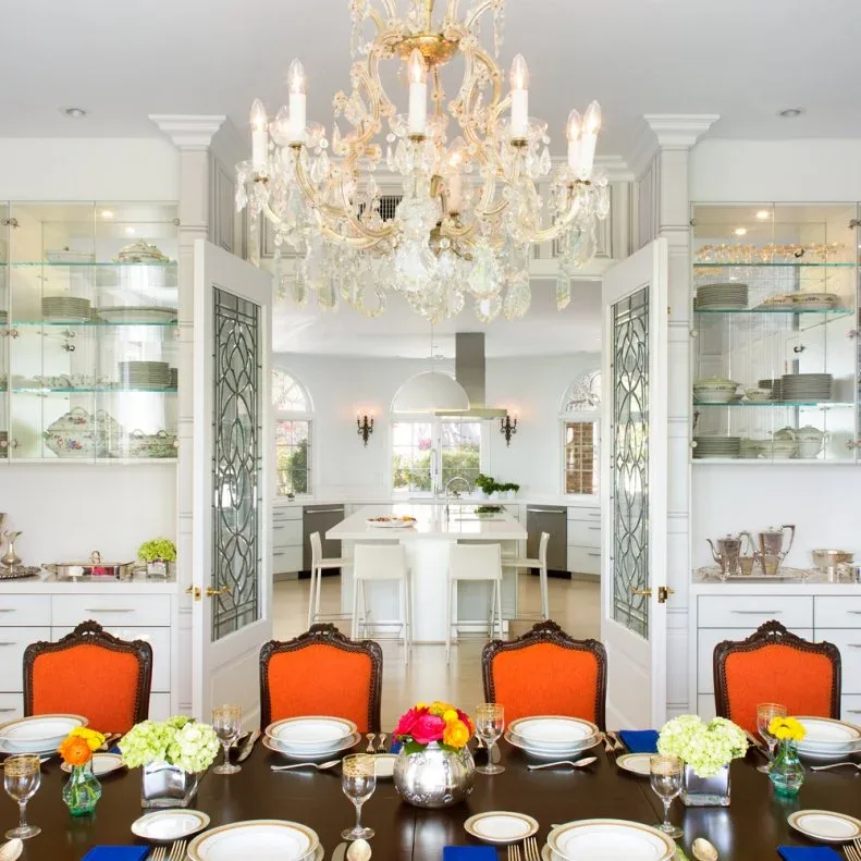 Dining room with an ornate gold-colored chandelier, orange-backed carved chairs, dark table and white glass-front dish displays