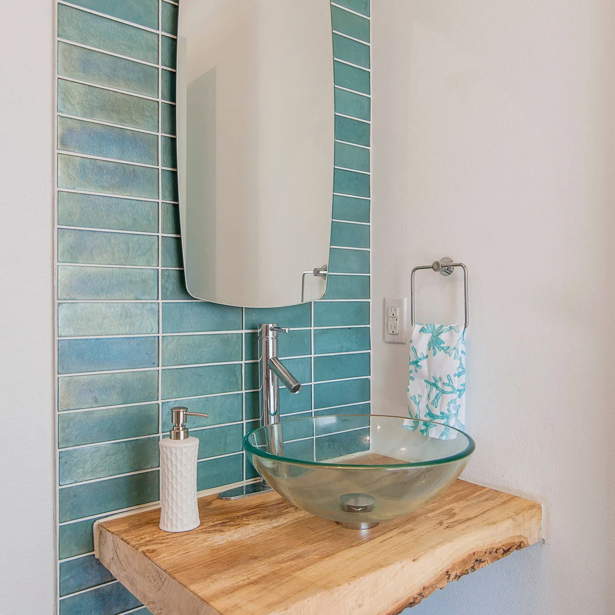 A clear glass vessel bowl rests on a wood counter with an irregular front edge against blue rectangular tiles and a slim mirror.