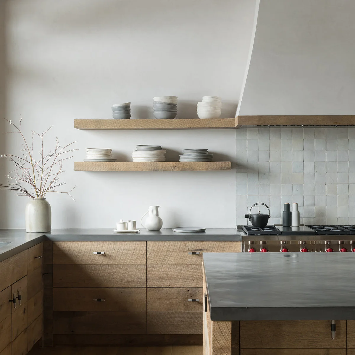 Pale wood drawers and shelves sit below gray counters beside a pale tiled cooking wall and a broad pale hood.