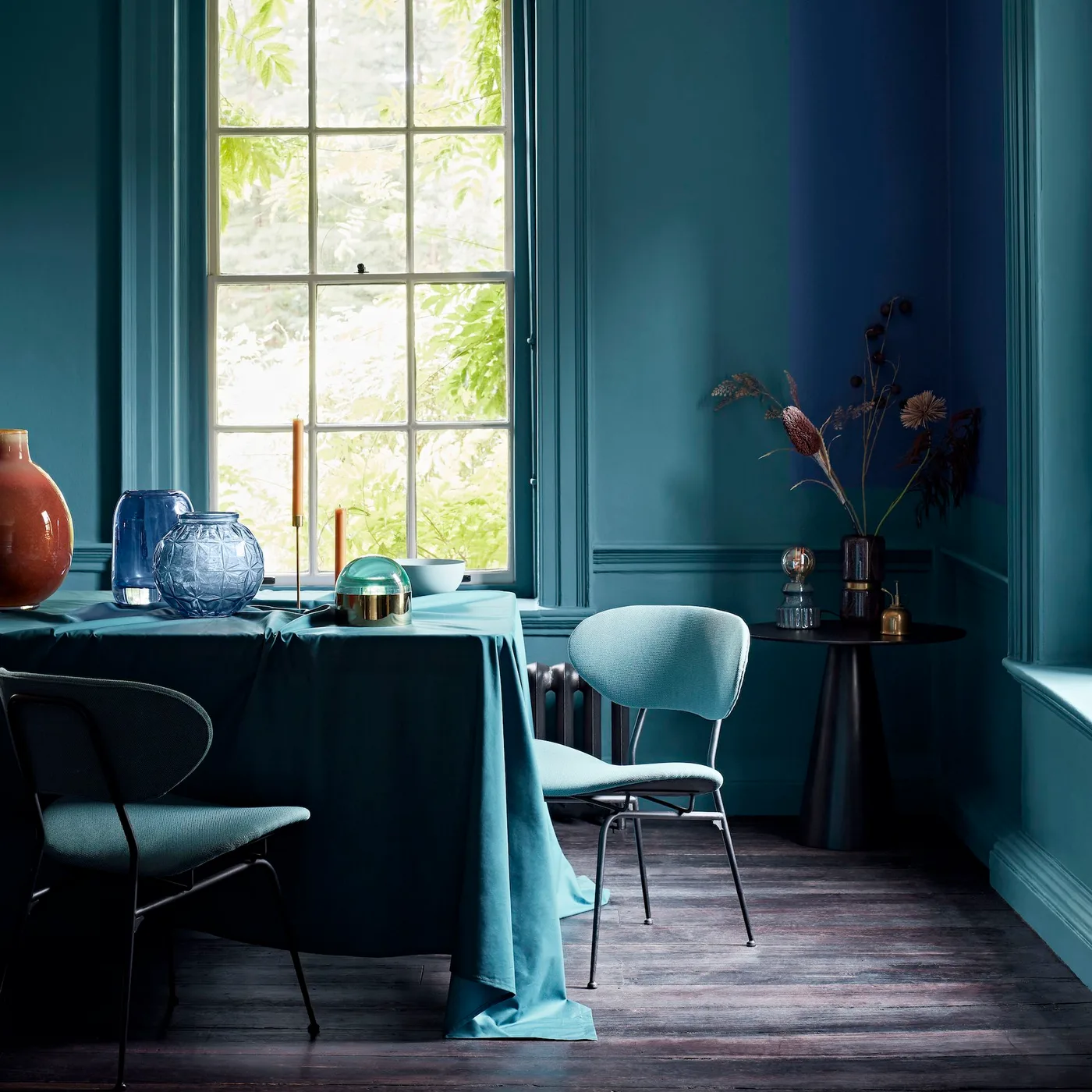 Deep teal paneled walls surround a cloth-covered dining table with pale blue chairs, colored glassware and slim candles beside a tall window