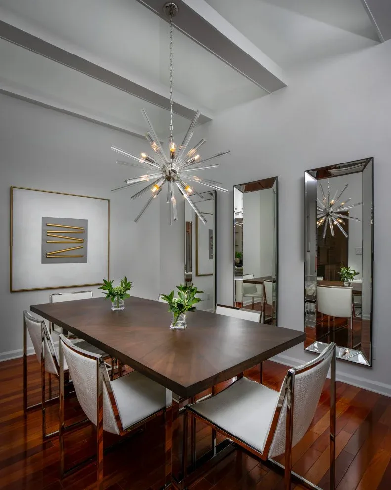 Wooden rectangular dining table with pale metal-framed chairs, three tall beveled mirrors and a clear starburst chandelier.