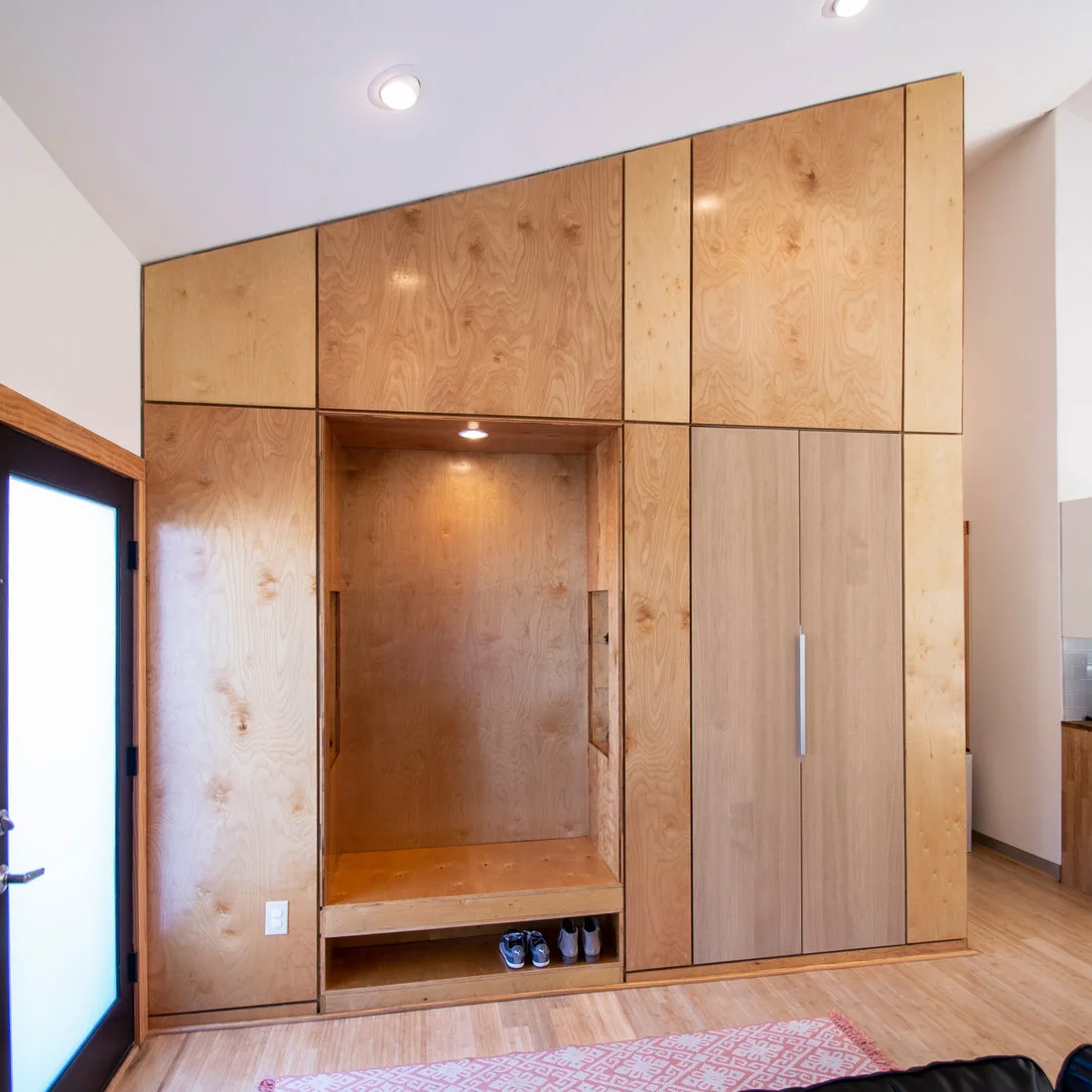 A full-height wood storage wall contains a recessed bench, lower shoe opening and tall cupboard beside a glazed entry door.