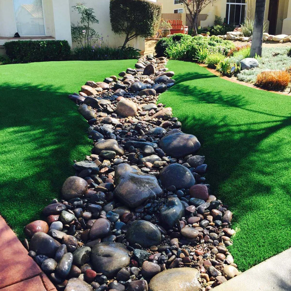 A curving channel of mixed river stones runs between bright synthetic turf and planted beds