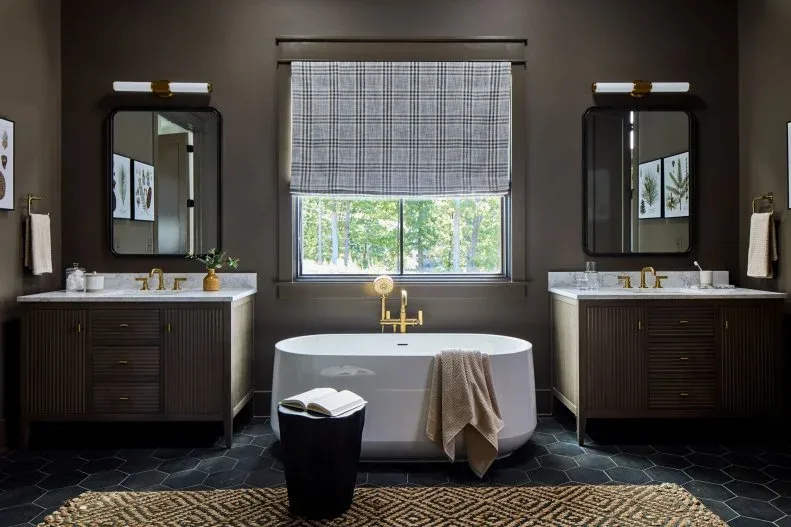 Deep gray bathroom with two dark fluted vanities, white central tub, black mirrors and picture lights, patterned blind, and charcoal hexagonal floor.