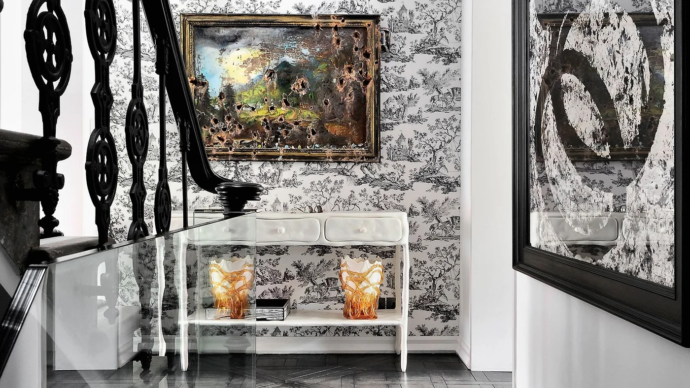 A framed colorful painting hangs on black-and-white scenic wallpaper behind a white console with amber objects beside an ornate black railing