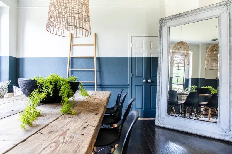 Large pale floor mirror beside a long wood table with black chairs and two-tone blue and white walls