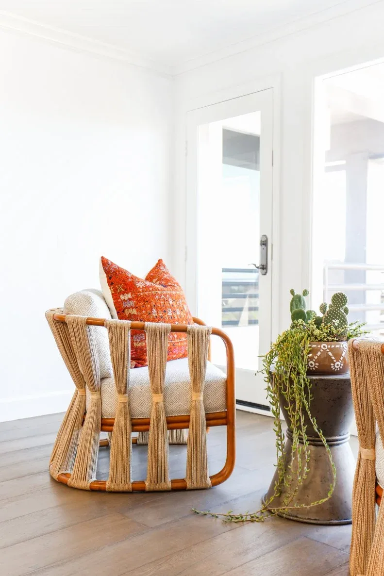 An open wood-and-cord chair has a pale seat and orange patterned cushion, with a trailing plant on a metal stand beside a bright doorway.