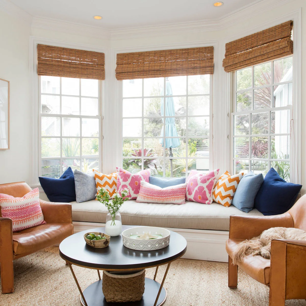 A pale bay-window seat with pink, orange and blue pillows, woven blinds and two brown armchairs.