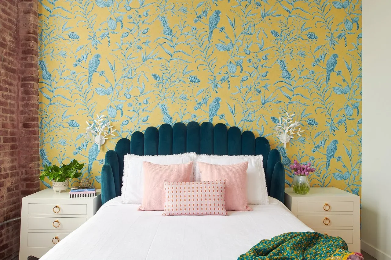 Yellow wallpaper with blue birds and foliage frames a blue scalloped headboard, white bedding, pink cushions and pale drawer tables.