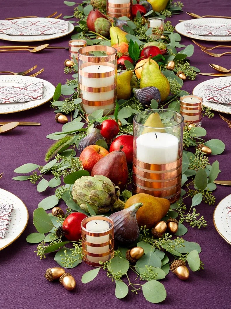 Green leaves, pears, artichokes and red fruit form a runner on plum cloth beside white patterned plates and striped candleholders