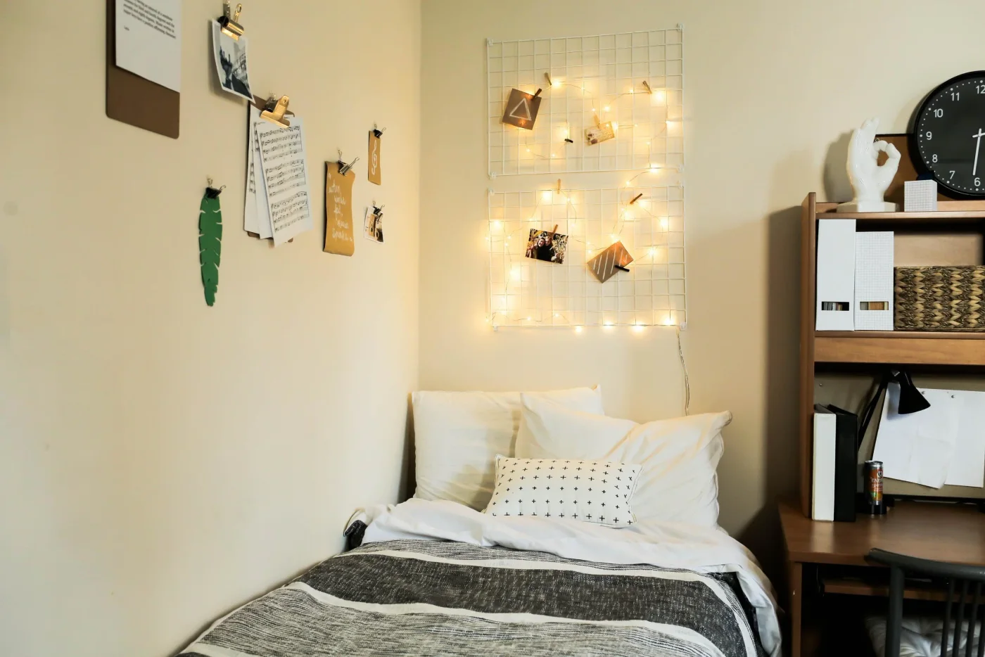 Student bed with black-and-white throw beneath two wire grids holding photos and warm string lights