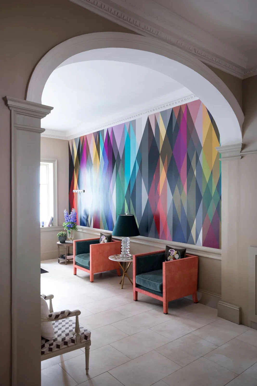 A multicolored geometric wall stands behind orange-framed chairs with dark teal cushions and a small lamp table beneath a pale arch