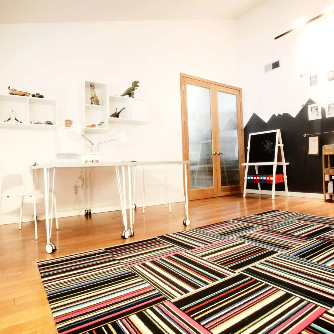 A white playroom has two white tables on casters, open display shelves, a black mountain-shaped chalkboard wall, a wood-framed glazed door and a striped rug.