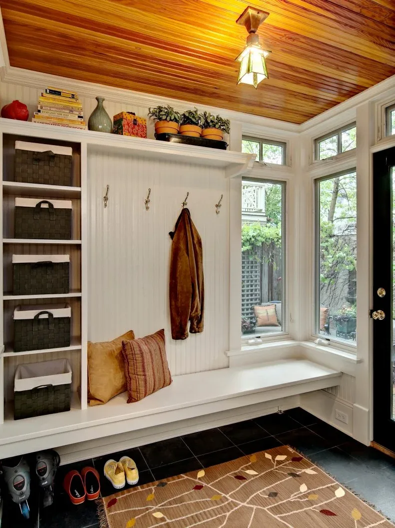 A long white bench extends beneath coat hooks and corner windows, with basket shelves at one end and shoes on the floor below.
