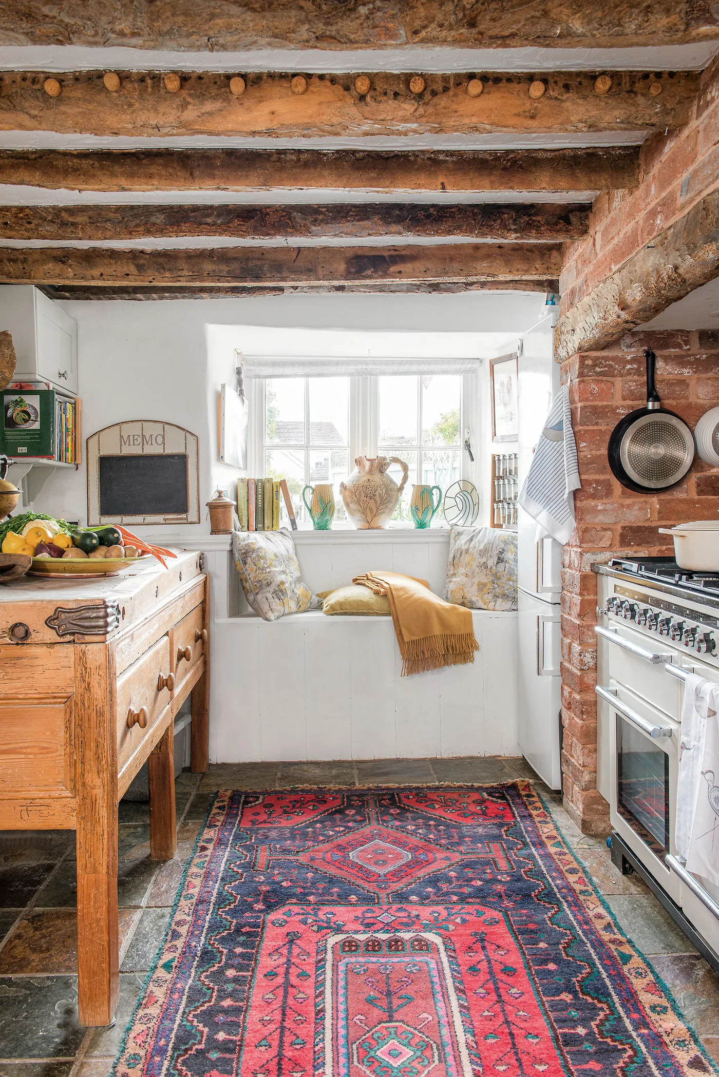 Rustic kitchen aisle with a red-and-blue patterned rug, exposed brick, ceiling beams and a window bench.