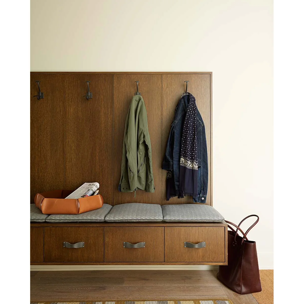 Wall-mounted wooden entry bench with three drawers, gray seat pads and coat hooks on a wood back panel.