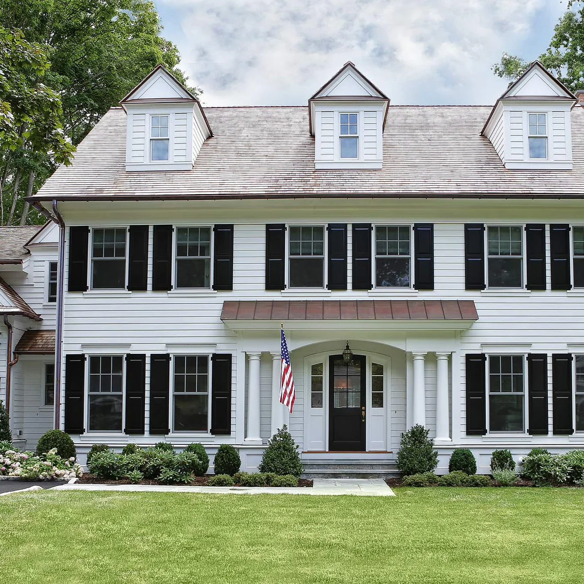 A white Colonial house with black shutters, paired upright shrubs near the entrance and lower green planting beneath the windows.