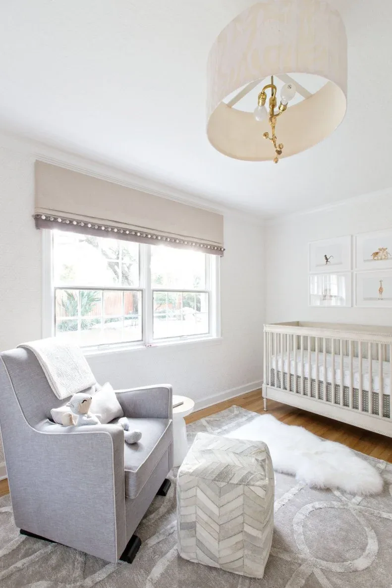 A gray adult chair, patterned pouf and pale crib sit beside a window in a soft gray nursery