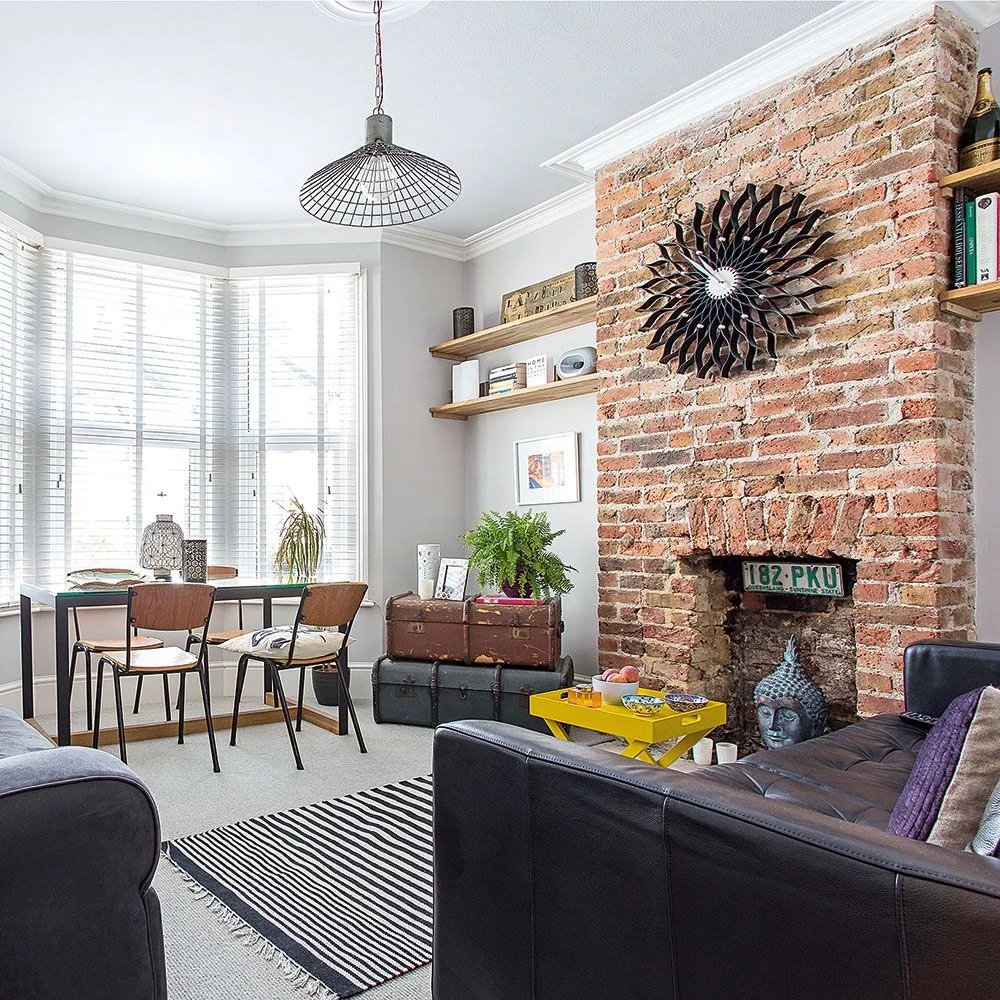 A brick chimney wall stands beside wooden shelves, dark seating and a small dining table under pale walls