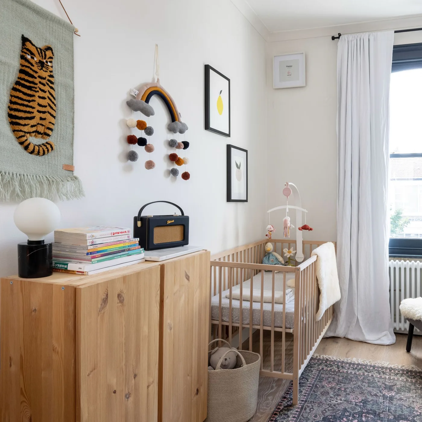 Wooden crib and dresser in pale nursery with tiger wall textile, rainbow hanging, framed pictures, white curtains and dark patterned rug.