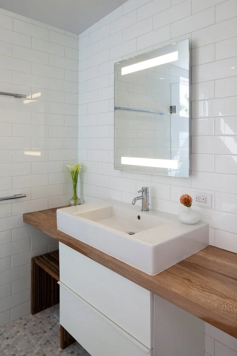 A white rectangular vessel basin sits on a wood counter below a lit rectangular mirror beside white subway tiles and a clear shower screen