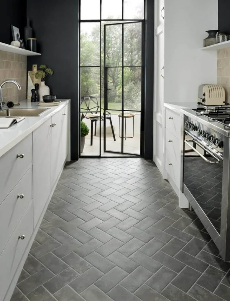 A narrow white kitchen has a gray herringbone tile floor, dark upper wall sections, pale counters and a black-framed glazed door at the far end.
