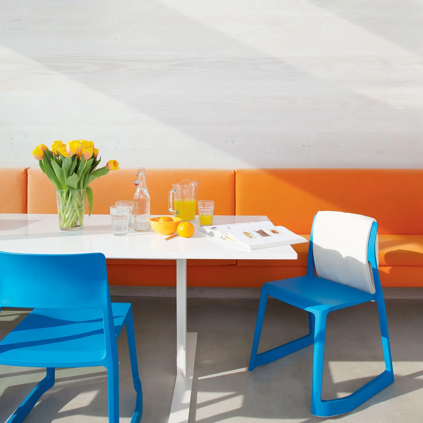 Dining area with a long orange upholstered banquette, white table, blue and white chairs and pale surrounding walls.