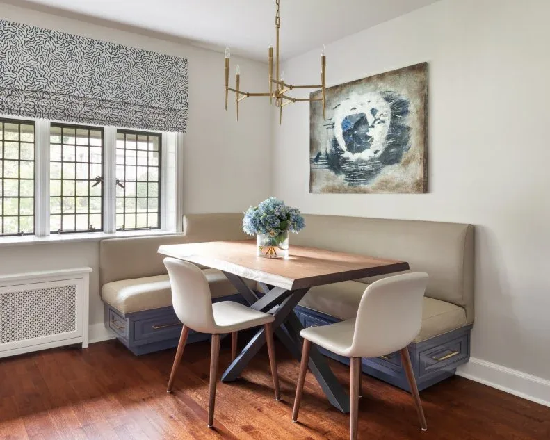 Pale upholstered corner bench with blue-gray drawers beneath the seats, a wooden trestle-style table, two pale chairs, patterned window shade, and abstract art.