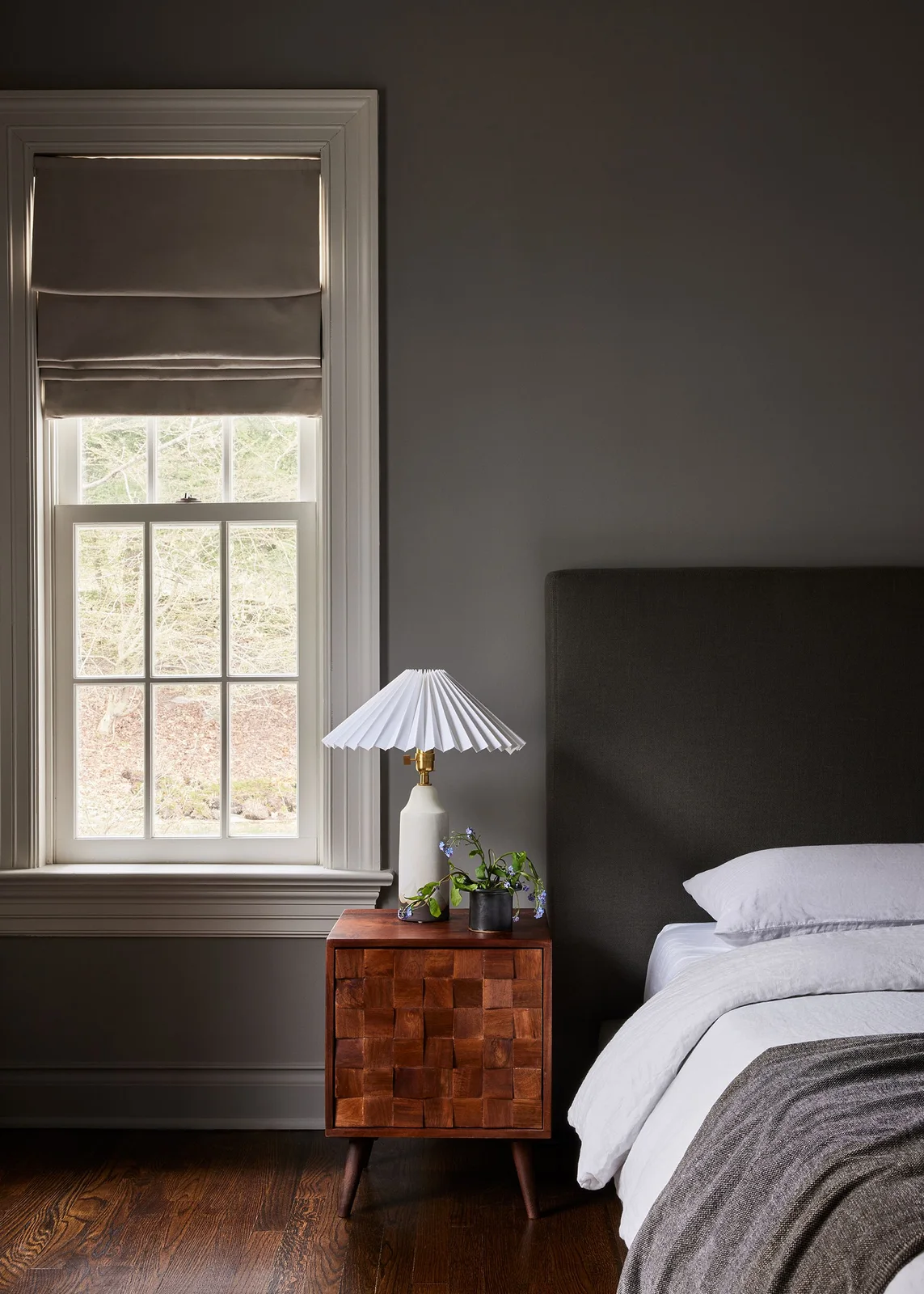 A dark gray bedroom wall frames a pale bed, a small wood cabinet with a block-patterned front and a white pleated lamp beside a sash window.