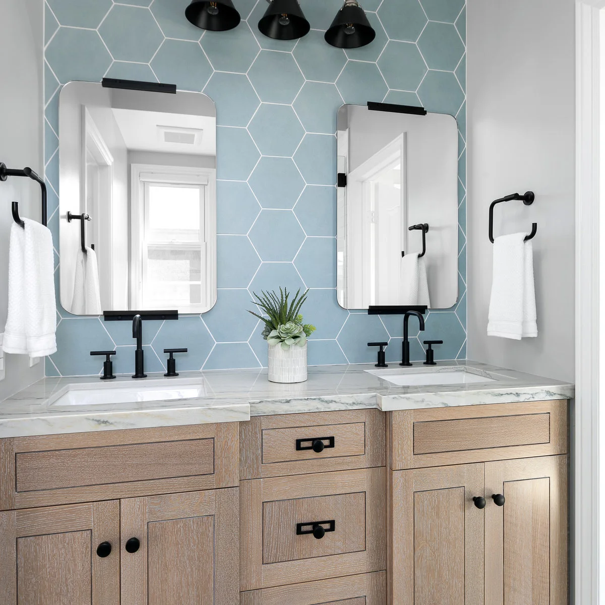 Light wood double vanity with blue hexagonal wall tiles and one white fluted pot of green foliage.