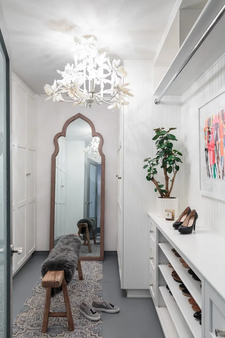 A slim white dressing closet has low shoe shelves, a narrow bench, a tall mirror and a floral ceiling light.