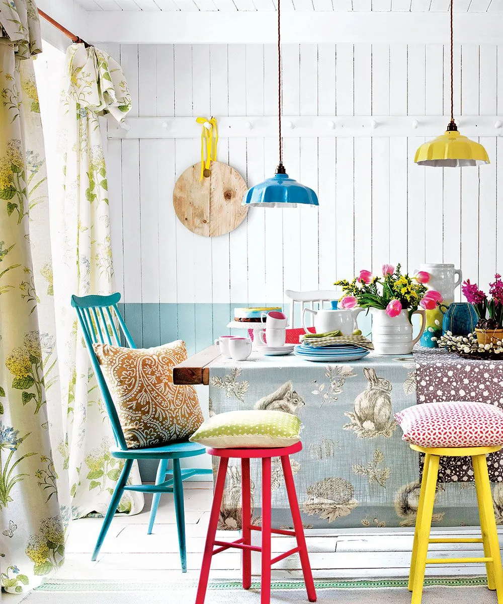 Pale botanical curtain at a dining-room window beside white and blue wall boards, a teal chair, red and yellow stools, patterned table coverings, and colored lights.