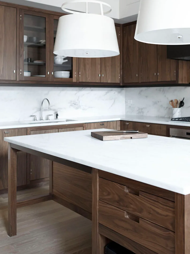 Dark brown kitchen with glazed upper cabinets, pale veined backsplash, white island counter, white pendant shades, and wood floor.