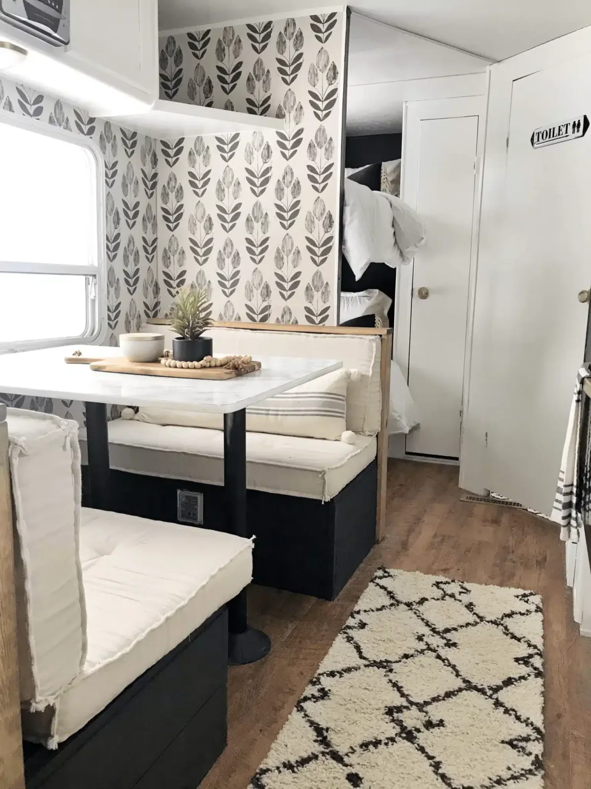 Camper dinette with white cushions, dark bench bases and a black-and-white leaf-patterned wall.
