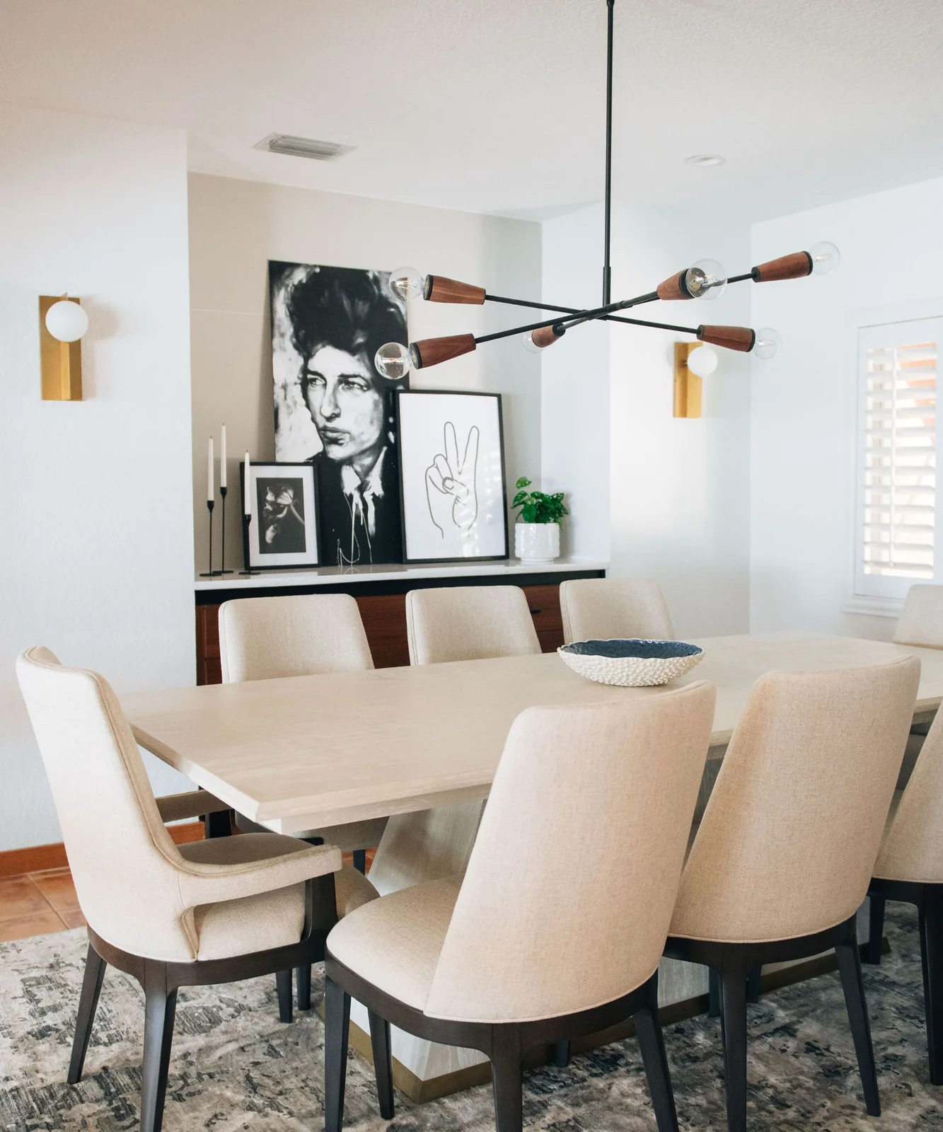 Pale upholstered dining chairs surround a pale table beneath a branching light with wood-colored ends beside framed black-and-white art and a two-tone sideboard