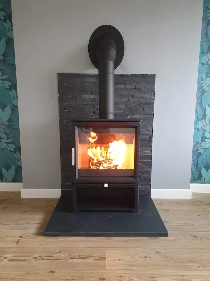 A black stove with an open lower compartment sits on a square slate hearth against a textured dark wall.