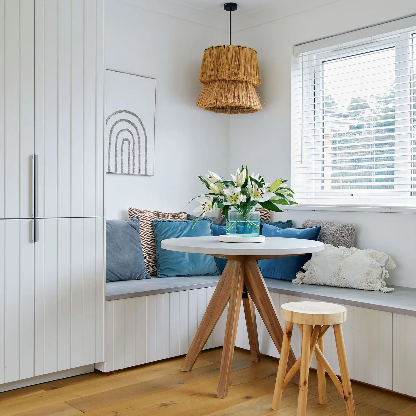 Window dining nook with an L-shaped cushioned bench, blue-gray pillows, round pale table, wooden stool and woven pendant.
